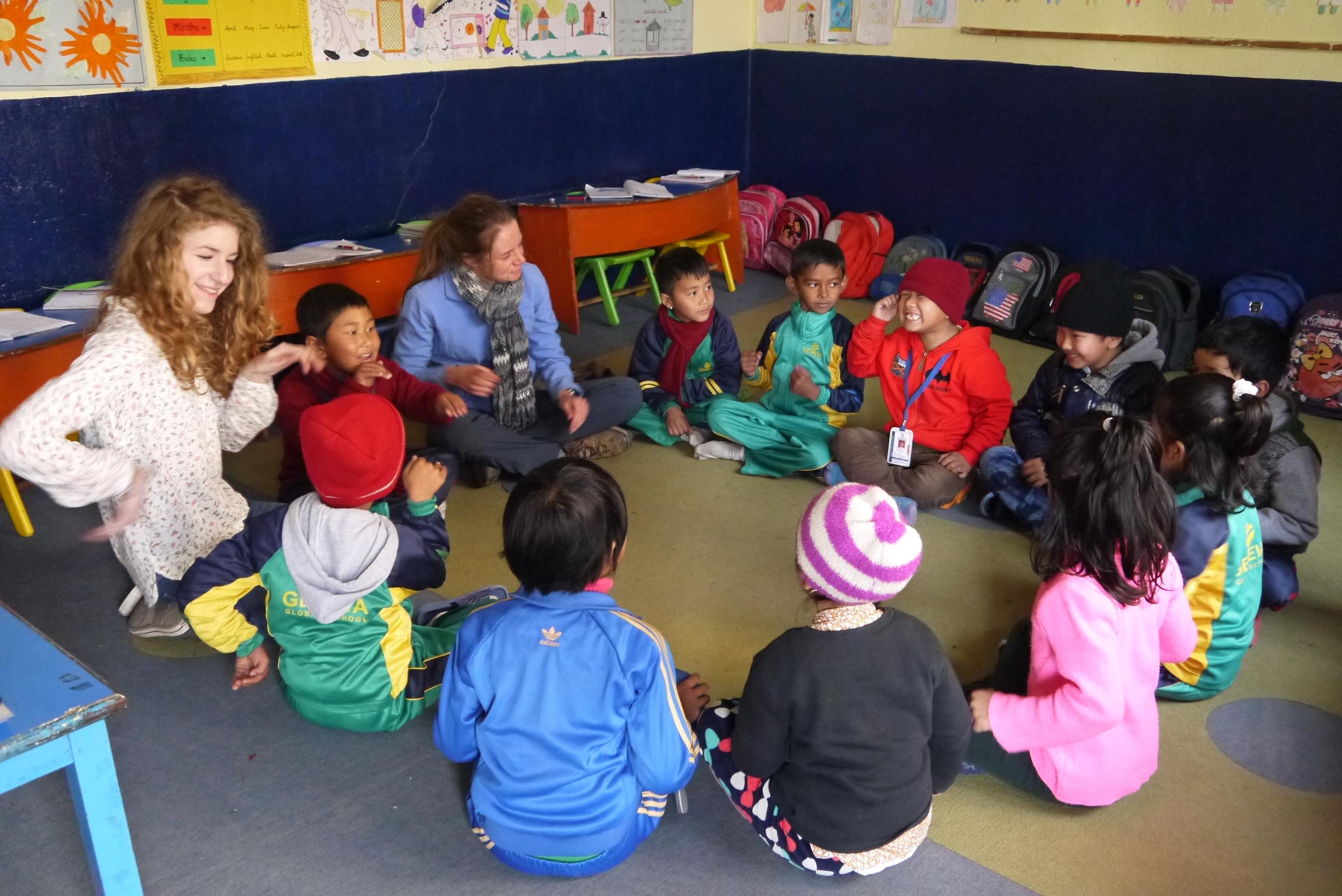 Female volunteers sit with children in a circle during an educational activity at a Childcare placement in Nepal. Projects Abroad Childcare volunteers in Nepal sit down with children and play educational games ar a kindergarten.